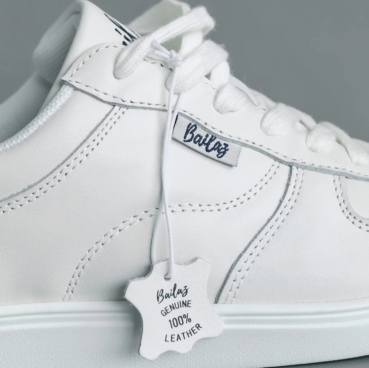 Close-up of a white dance shoe with the brand name 'Bailaz' on the lace tag, featuring a genuine leather upper and a proprietary polyurethane composite outsole.